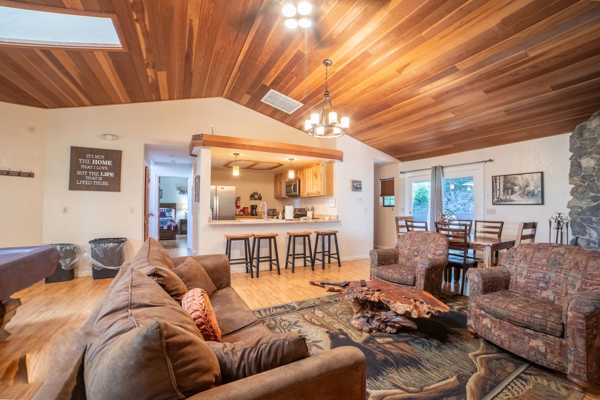 Cozy cabin interior: living area with brown sofa, bar stools, kitchen, dining table, wooden ceiling, and fireplace.