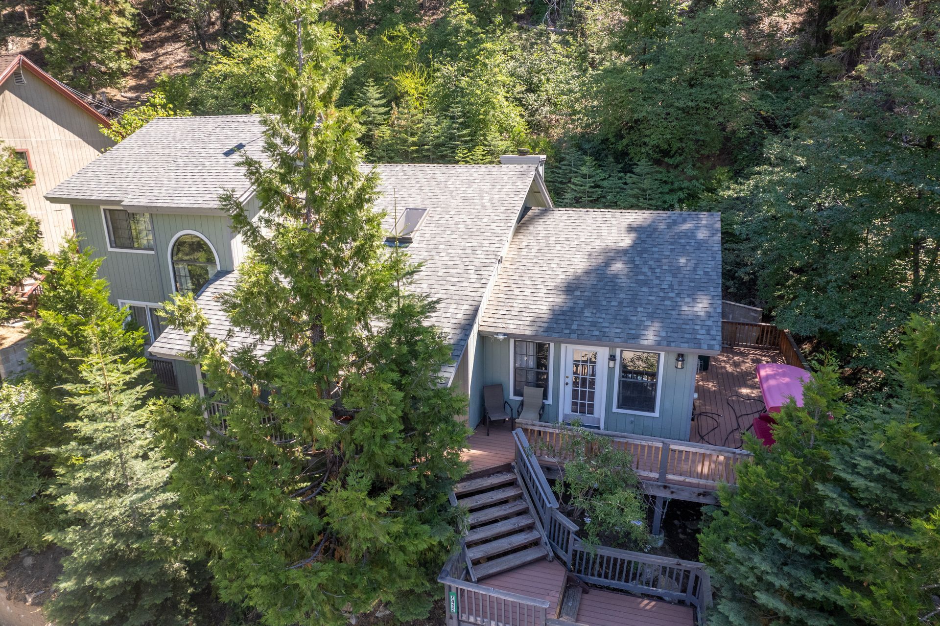 Two-story house surrounded by trees, with a wooden deck and stairs. Gray roof and light-blue exterior.
