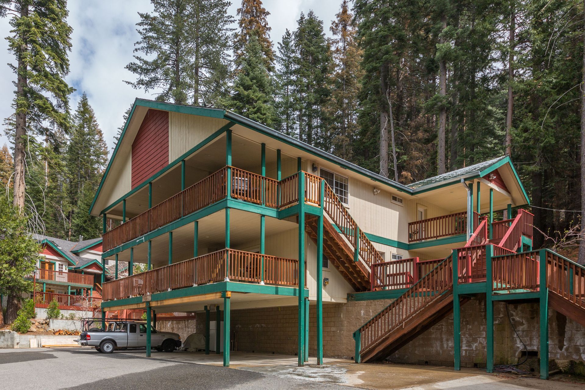 Two-story building with wooden balconies. Green trim, brown railings, and stairs. White truck parked below.