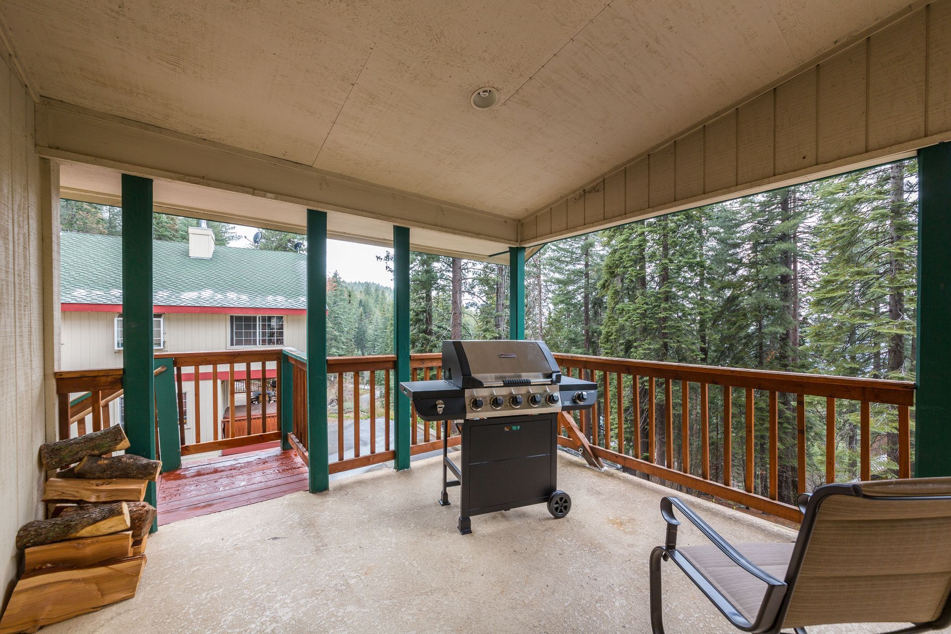 Covered porch with grill, firewood, and chair, overlooking a forest and house.