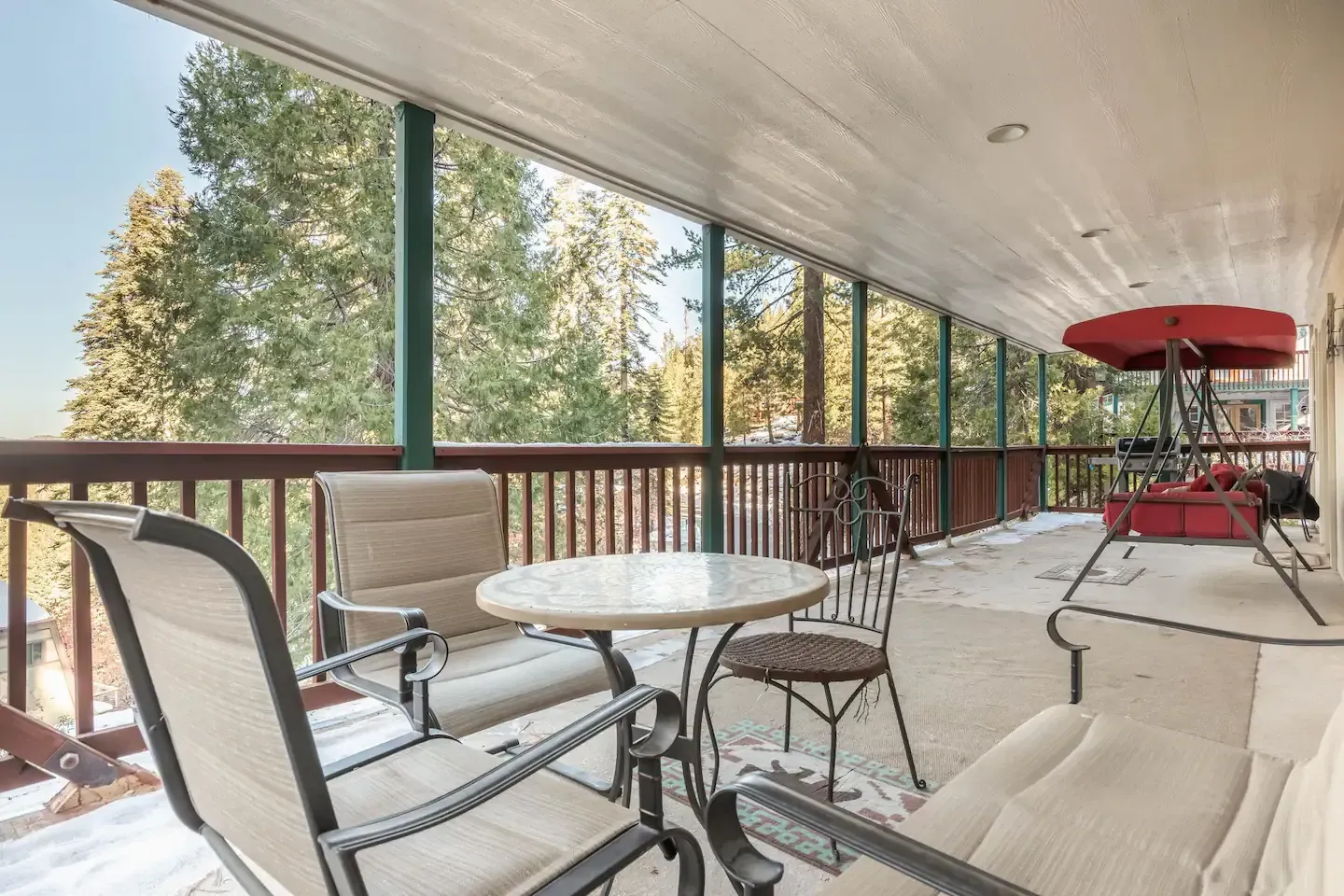 Patio with seating overlooking a snowy forest. Red umbrella, brown railing, and green trees.