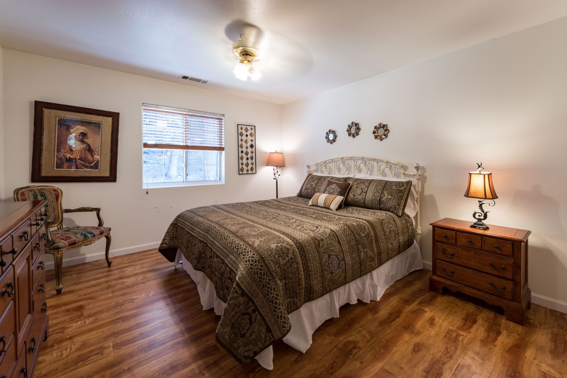 Bedroom with a bed, dresser, chair, and nightstand with lamps. Wooden floors, white walls, and a window.
