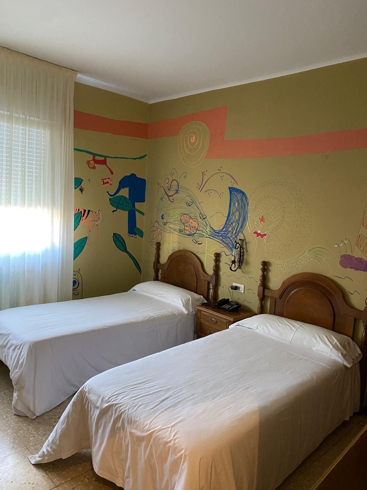 Dos camas individuales en una habitación colorida con murales pintados en las paredes y una ventana con cortinas transparentes.