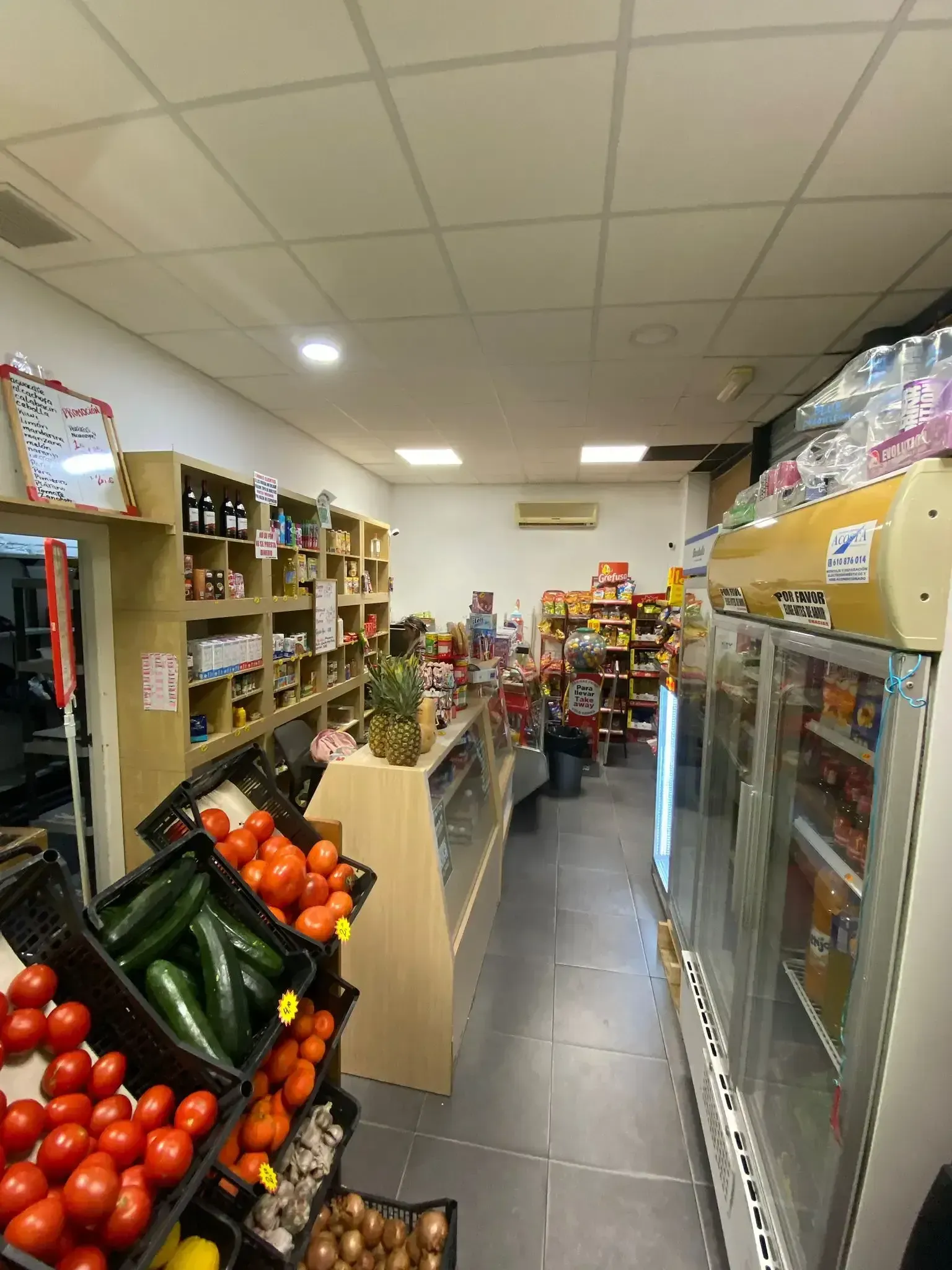 Pasillo estrecho de comestibles con exhibidores de frutas y verduras y vitrinas refrigeradas en un mercado pequeño.
