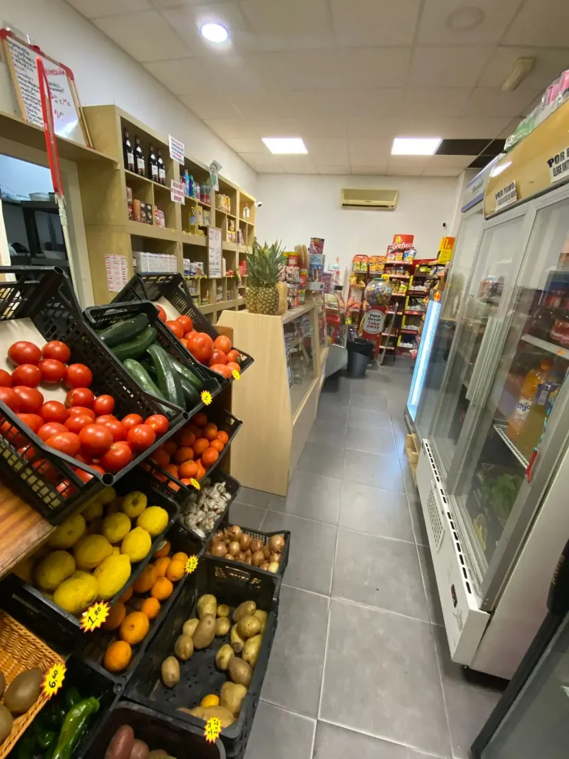 Pasillo estrecho de comestibles con exhibidores de frutas y verduras y vitrinas refrigeradas en un mercado pequeño.