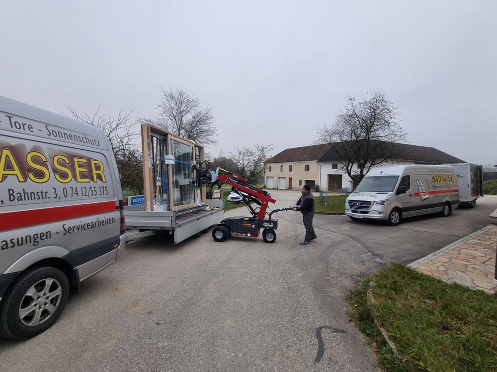 Arbeiter verwenden einen kleinen Kran, um ein großes Fenster von einem LKW abzuladen. Zwei weitere Lieferwagen stehen in einer ländlichen Umgebung.