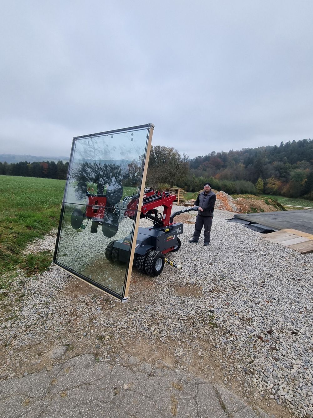 Ein Mann bedient eine kleine Maschine, die eine große Glasplatte auf einem Kiesweg im Freien transportiert. Bewölkter Himmel.