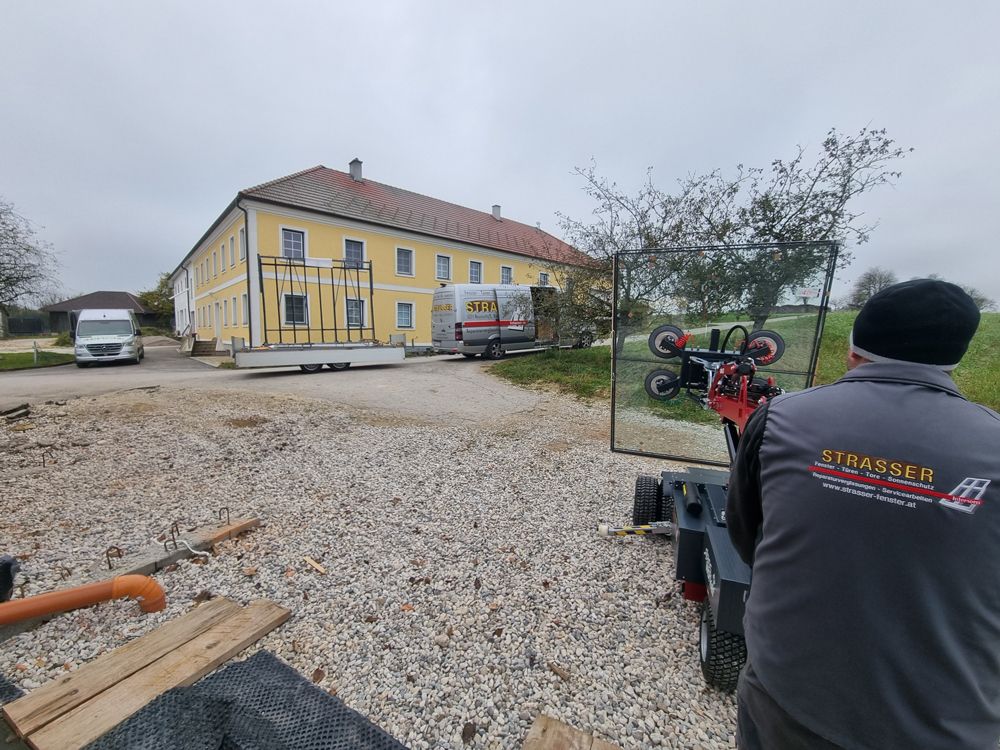 Person hält Glasscheibe vor einem gelben Gebäude mit Baumaschinen auf einem Schotterplatz.