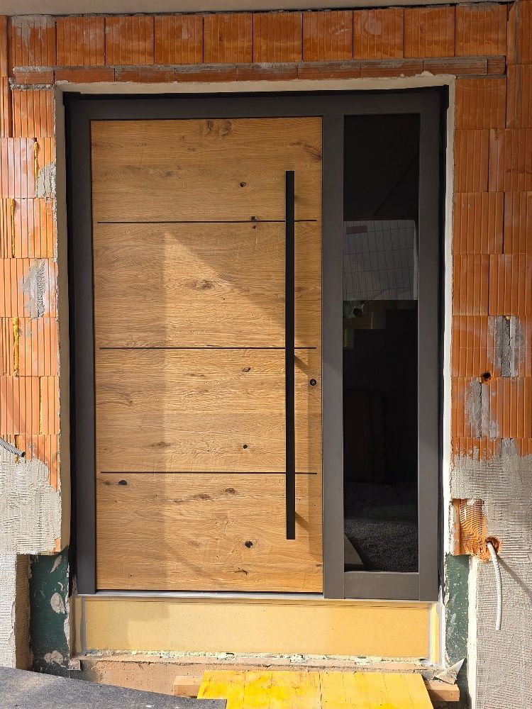 Eine moderne Haustür mit heller Eichenholzmaserung, einem vertikalen schwarzen Türgriff und einer seitlichen Glasscheibe in einer unverputzten Wand.