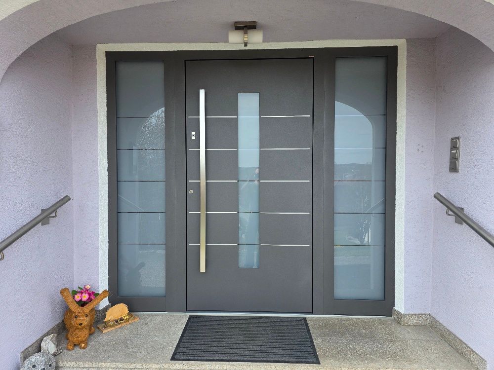 Eine moderne graue Haustür mit vertikalem silbernen Türgriff und Milchglasscheiben, die sich unter einem weißen, gewölbten Eingangsbereich befindet.