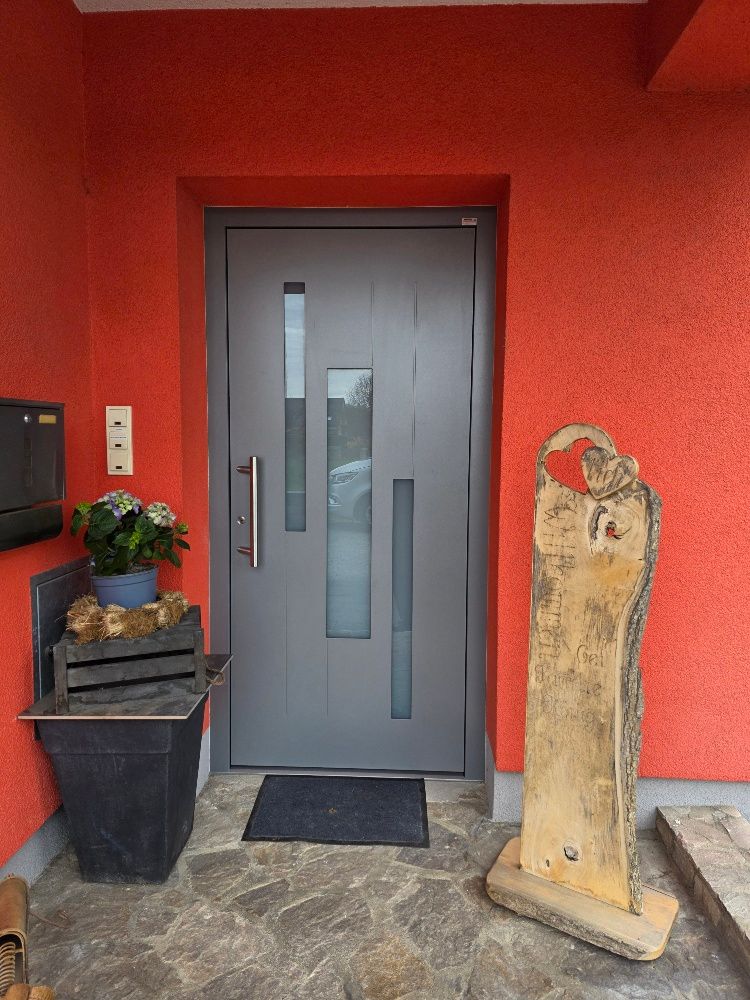 Eine graue Eingangstür mit vertikalen Glasscheiben vor einer strukturierten roten Wand mit einer Topfpflanze und einer Steinskulptur.
