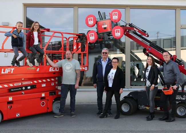 Menschengruppe bei roten Baumaschinen. Zwei Kinder auf Hebebühne, Erwachsene posieren bei Maschinen.