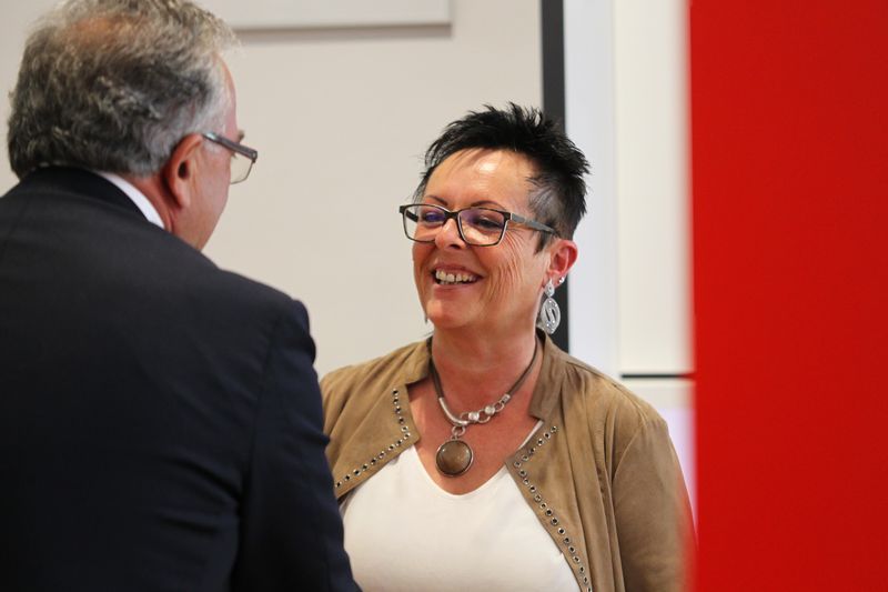 Ein Mann und eine Frau geben sich die Hand und lächeln. Die Frau trägt eine Brille und eine Halskette.