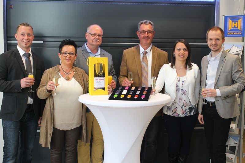 Eine Gruppe von Leuten steht mit Champagnergläsern in den Händen um einen Tisch herum.