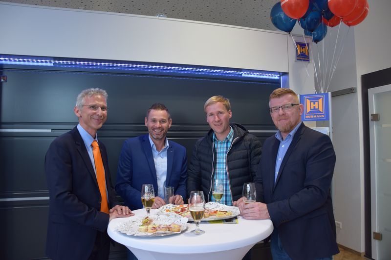 Eine Gruppe von Männern steht mit Tellern voller Essen und Gläsern Wein um einen Tisch herum.