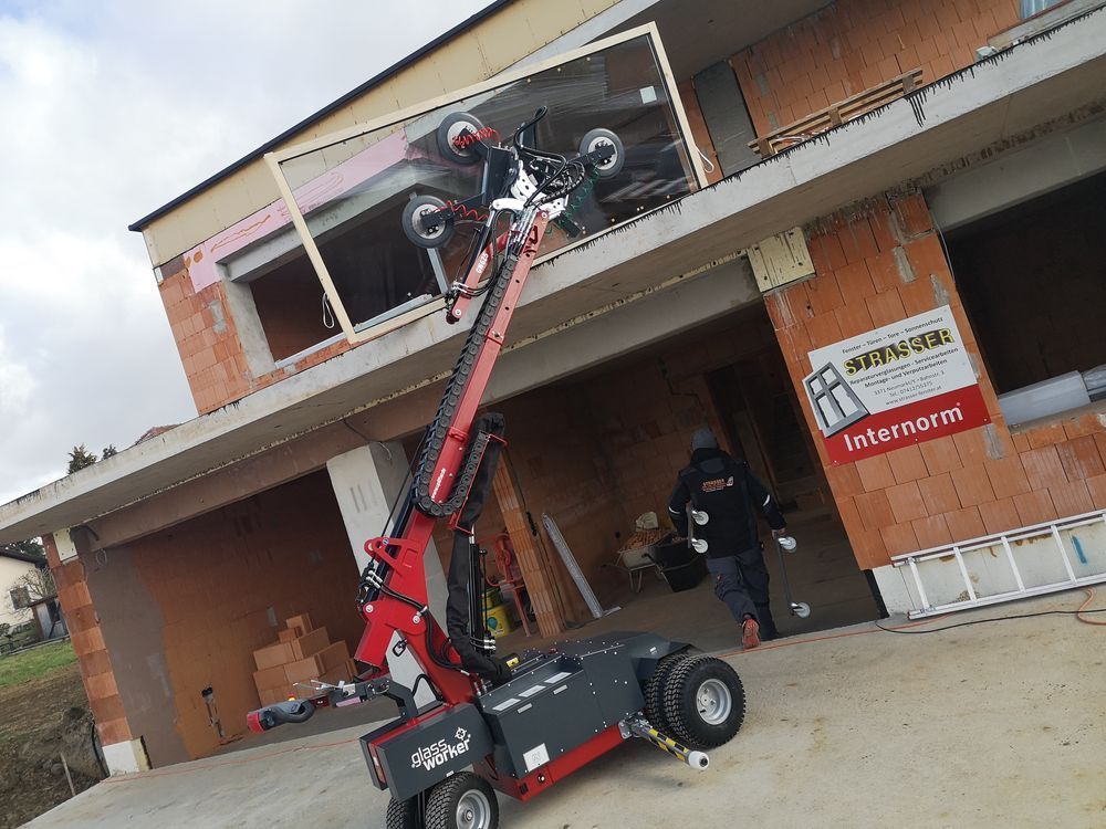 Eine rote Maschine mit Saugnäpfen hebt eine große Fensterscheibe in ein im Bau befindliches Gebäude.