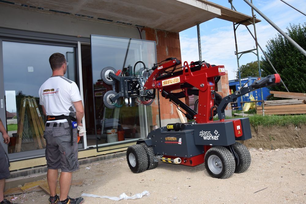 Ein Arbeiter beobachtet auf einer Baustelle einen roten Glasheberoboter, der Glas in einen Fensterrahmen einsetzt.
