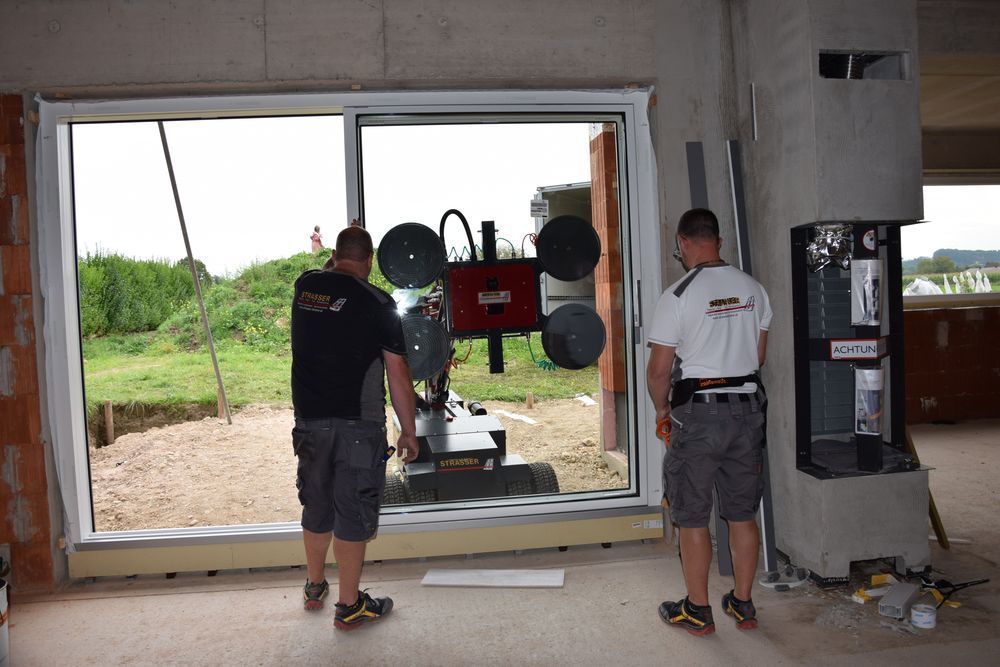 Zwei Arbeiter installieren auf einer Baustelle mit einem Saugheber ein großes Fenster.