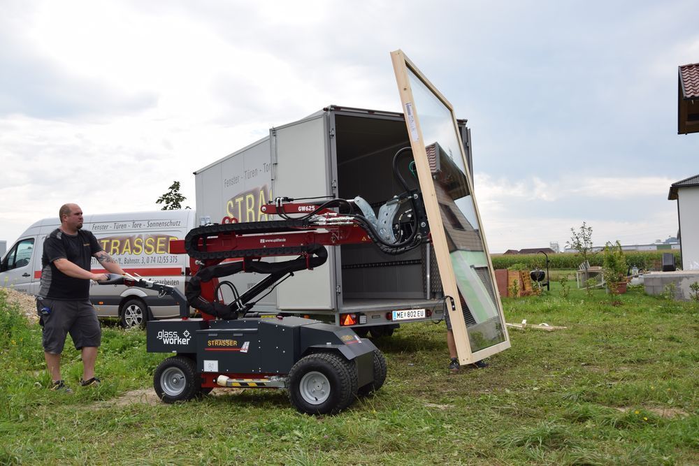 Mann bedient Roboterarm und lädt großes Fenster in LKW auf Wiese.