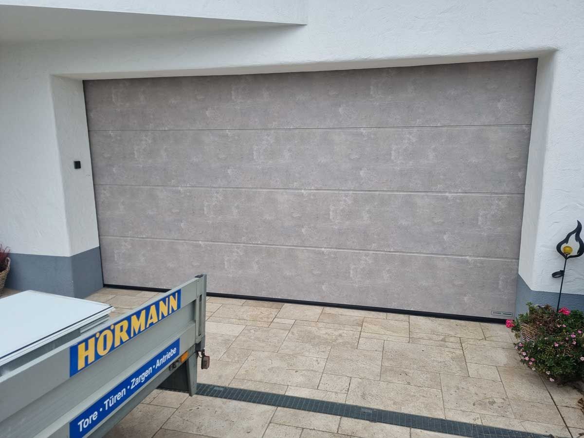 Geschlossenes graues Garagentor auf einer gepflasterten Auffahrt; davor steht ein kleiner Anhänger.