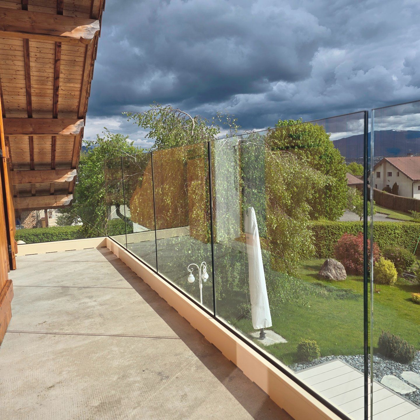 Balcon avec balustrade en verre donnant sur une cour verte, des arbres et des maisons sous un ciel nuageux.