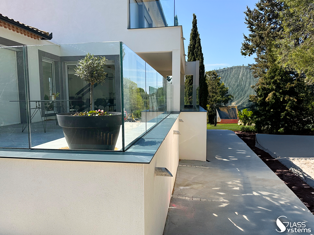 L'extérieur d'une maison moderne présente un balcon en verre transparent donnant sur un jardin avec une plante en pot.