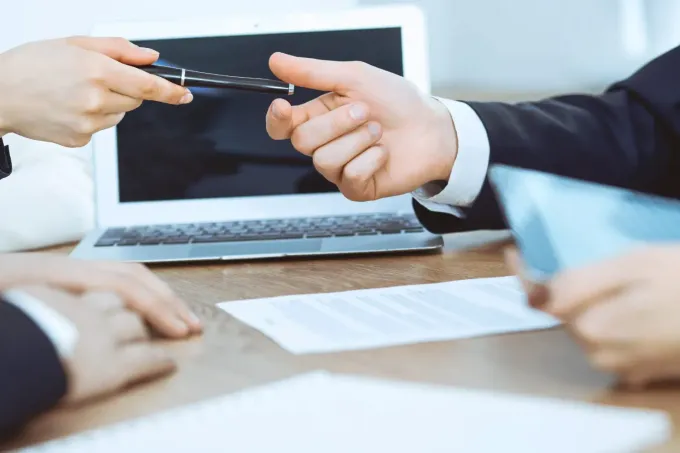 Dos profesionales sentados en un escritorio, pasándose una mano una pluma a la otra para firmar.