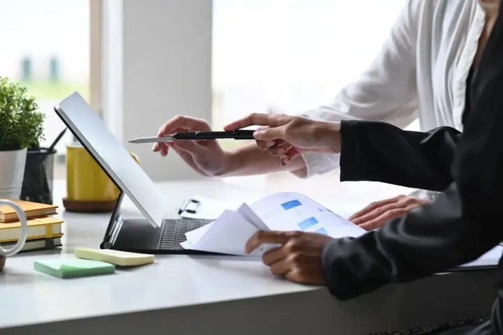 Dos personas, en un entorno de oficina luminoso, colaboran a través de la pantalla de un portátil.