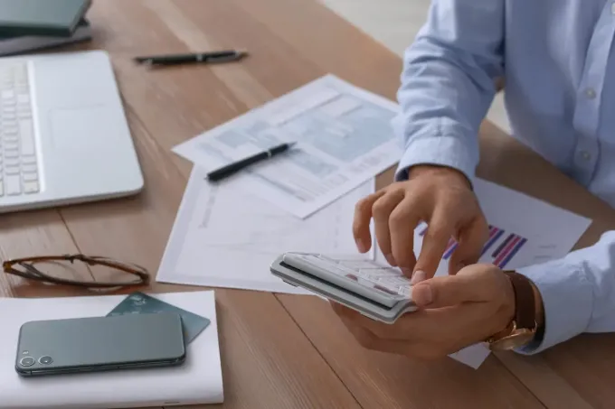 Una persona con una camisa azul claro trabaja en un escritorio, utilizando una calculadora sobre documentos financieros.