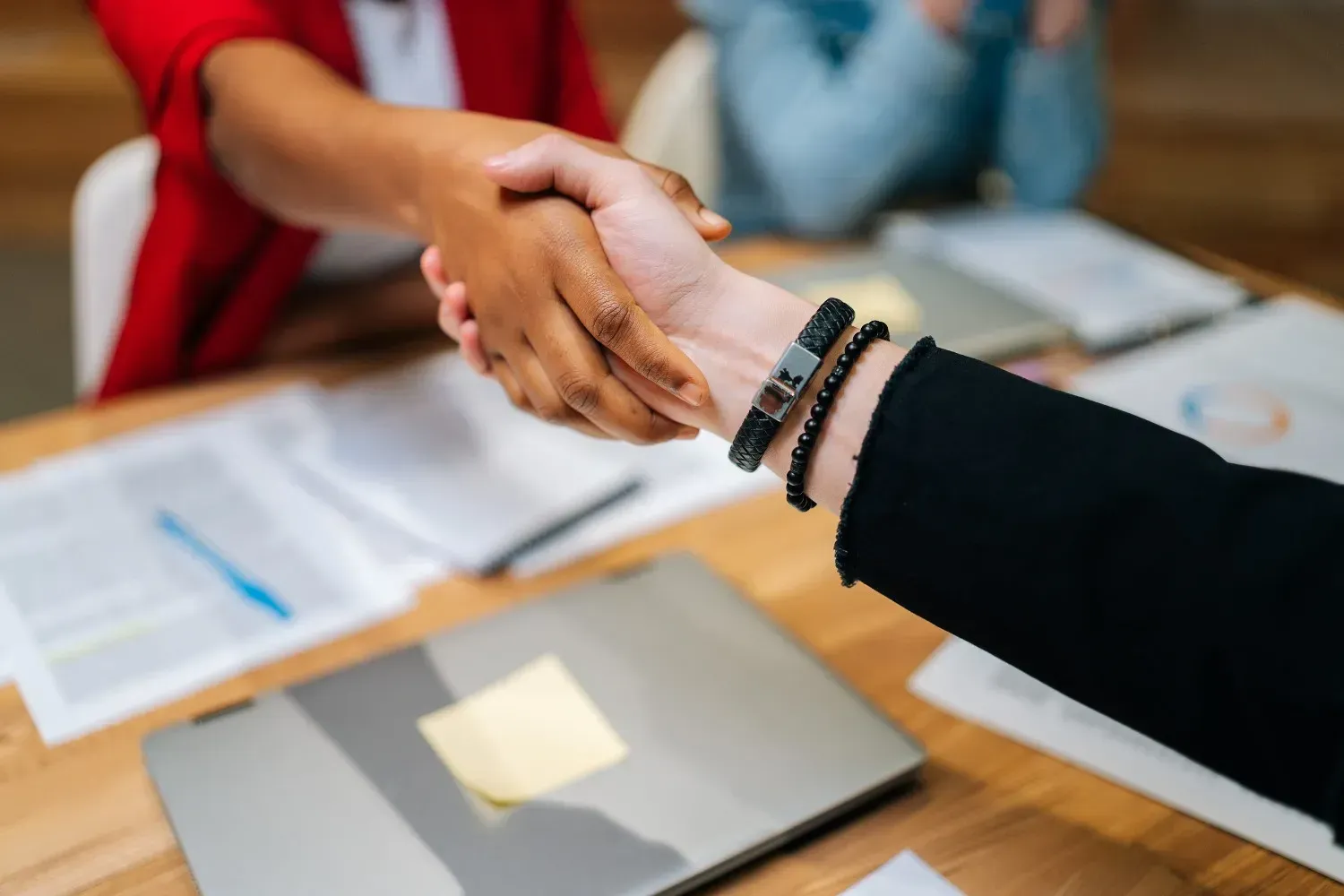 Dos personas dándose la mano sobre una mesa de madera con un ordenador portátil y documentos.
