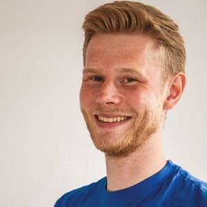 Ein Mann mit rötlich-blondem Haar und Bart lächelt vor einem weißen Hintergrund. Er trägt ein blaues Hemd.