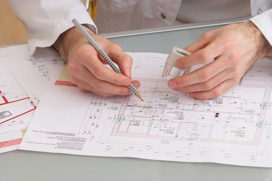Hände einer Person mit Bleistift und Radiergummi, die an Architekturplänen arbeitet und einen weißen Laborkittel trägt.
