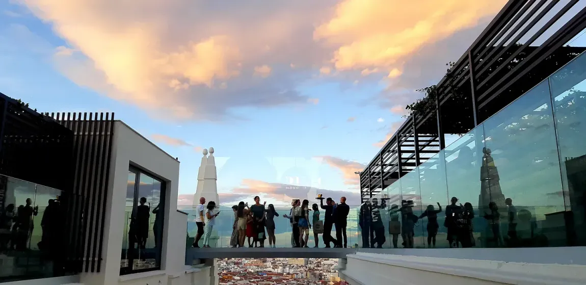 Gente en una terraza en la azotea viendo una puesta de sol sobre una ciudad.