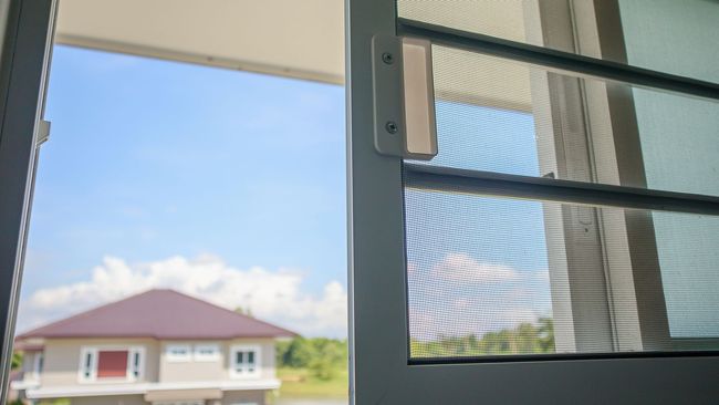 Vista a través de una ventana de una casa con techo rojo bajo un cielo azul; marco de ventana con mosquitera a la derecha.