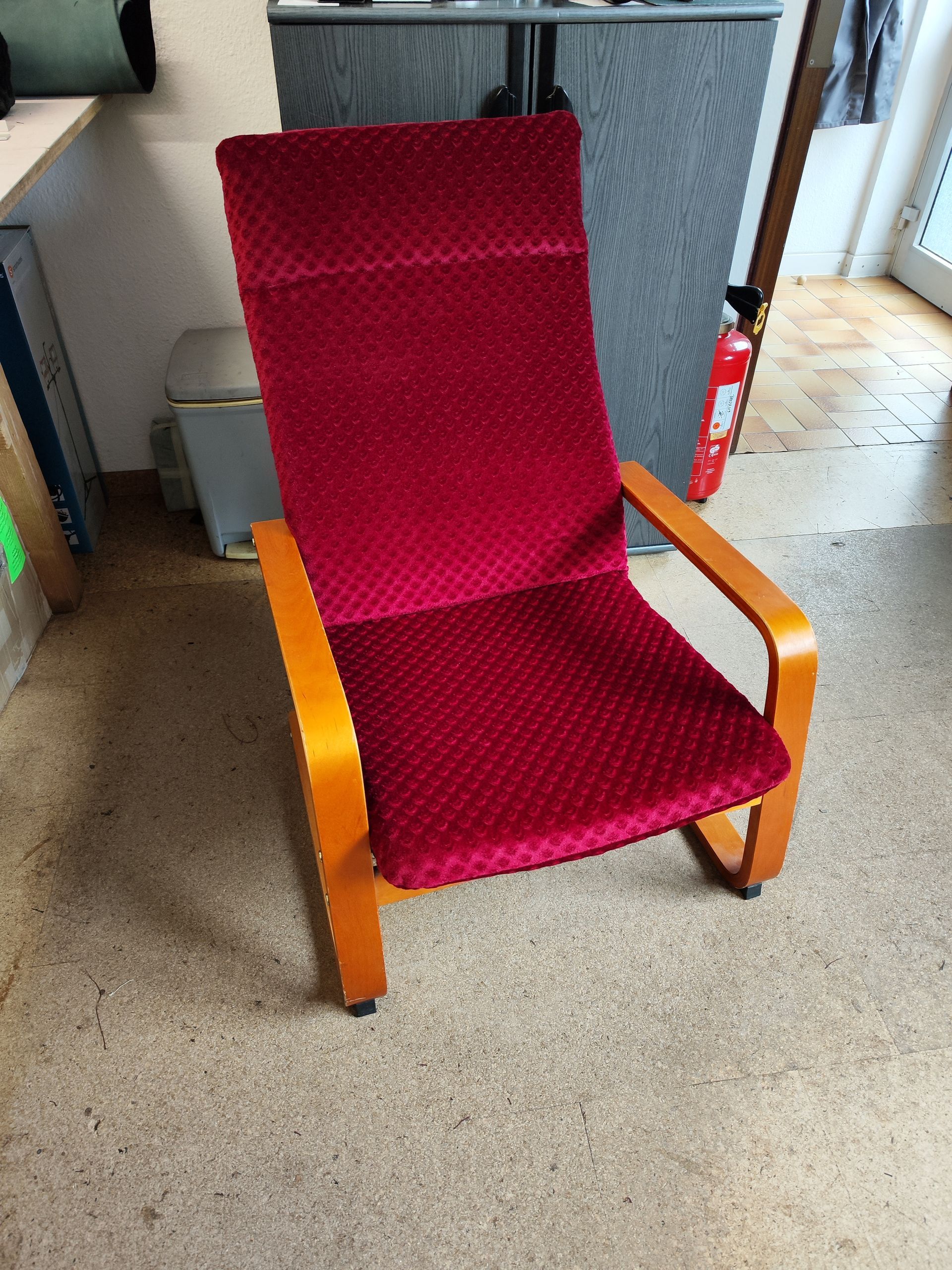 Auf einem Teppichboden steht ein roter Stuhl mit Armlehnen aus Holz.