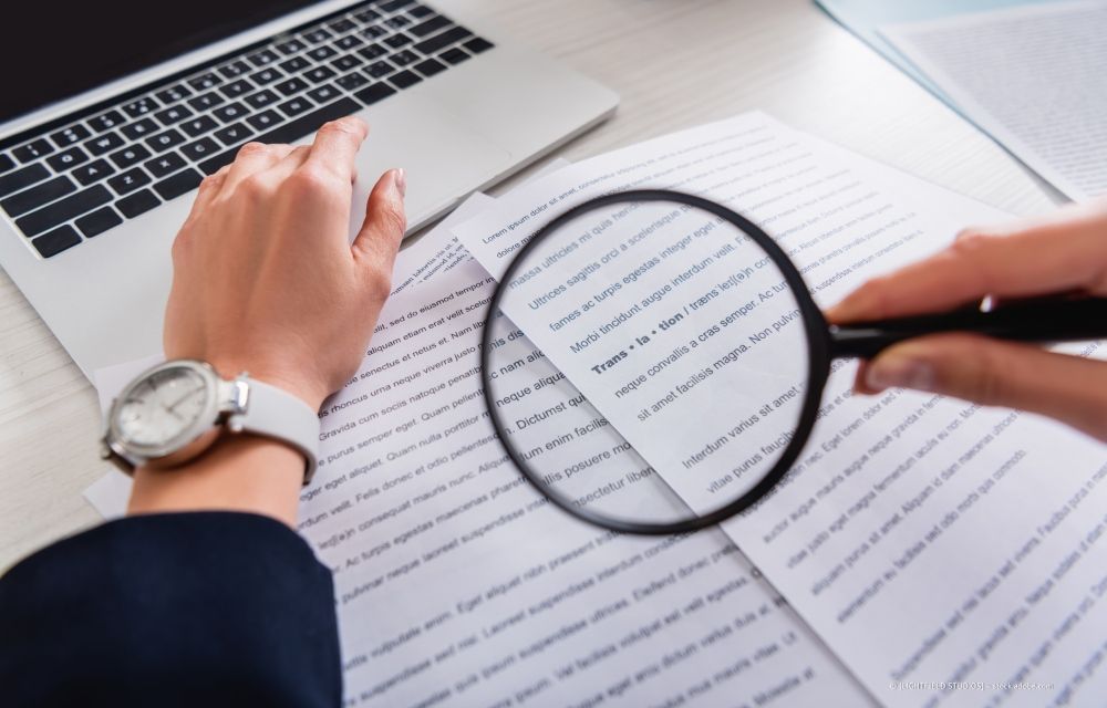 Eine Person benutzt eine Lupe, um neben einem Laptop einen Text auf Papier zu lesen.