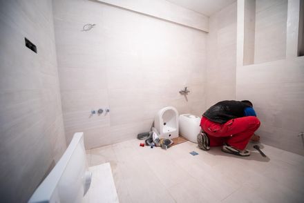 Baño sin terminar con azulejos blancos, inodoro, herramientas esparcidas y un carrito cubierto de rojo en el suelo.