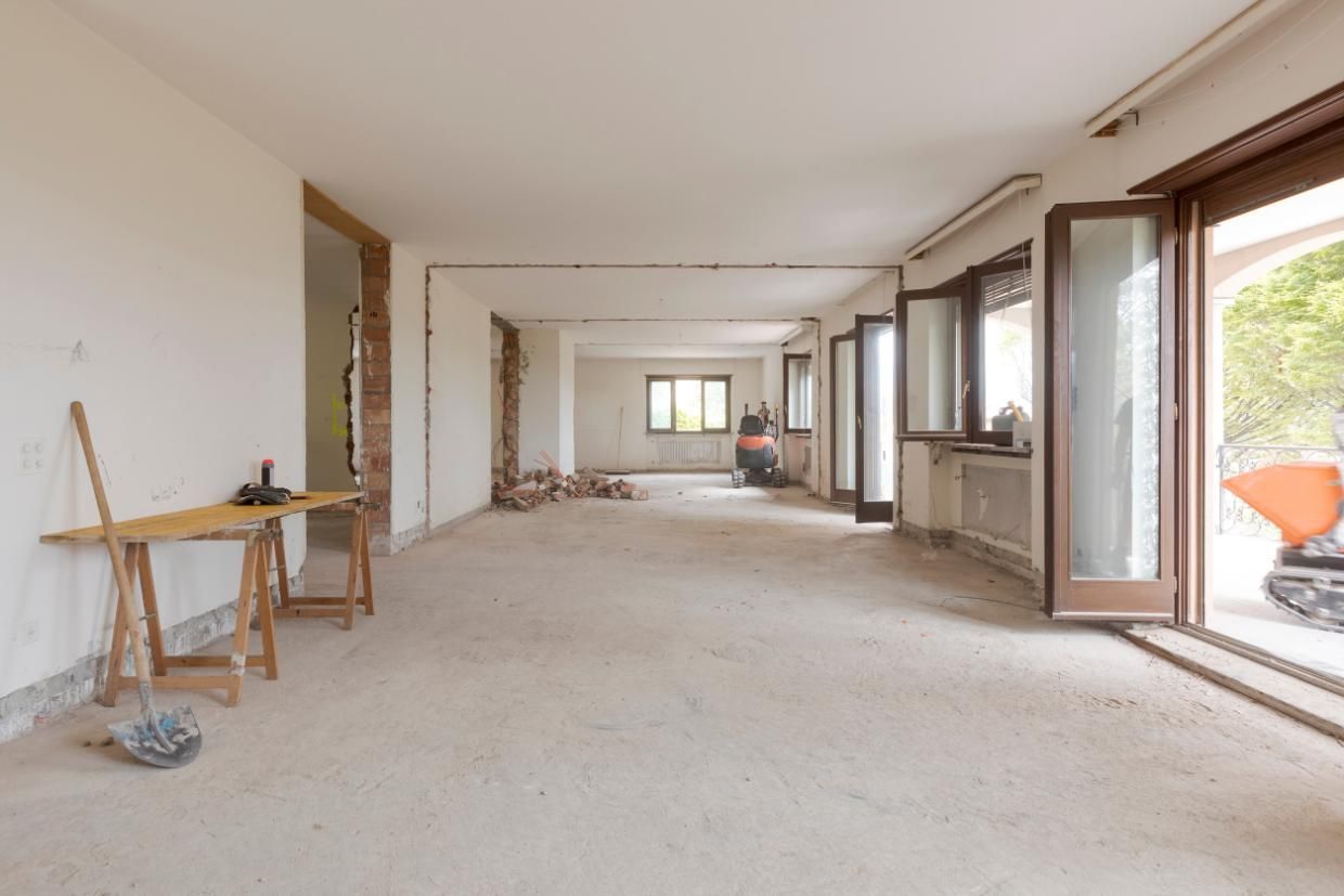 Habitación vacía en obras, con suelos desnudos, paredes blancas y ventanas y puertas abiertas.