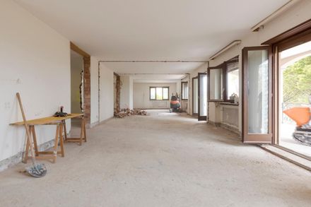 Habitación vacía en obras, con suelos desnudos, paredes blancas y ventanas y puertas abiertas.
