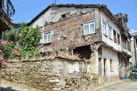 Antigua casa de piedra dañada, con paredes derruidas y un techo parcialmente derrumbado, en una calle soleada.