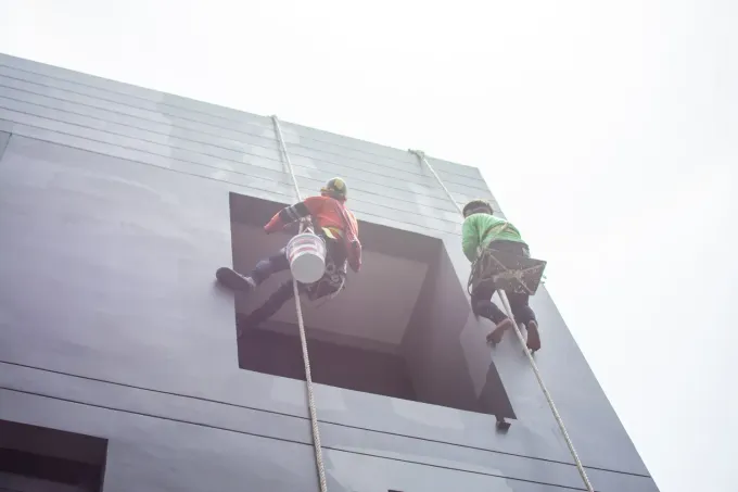 Dos trabajadores con trajes de seguridad rojos y verdes descienden en rápel por la fachada de un edificio.