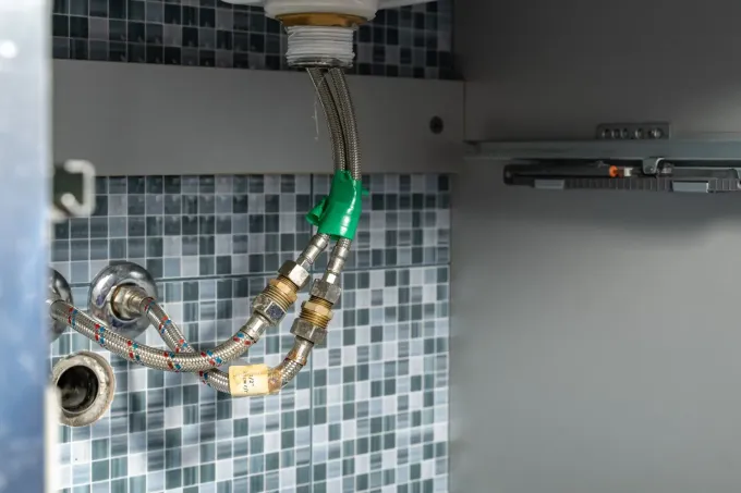 Instalación de fontanería bajo el fregadero con manguera trenzada y cinta verde contra una pared alicatada.