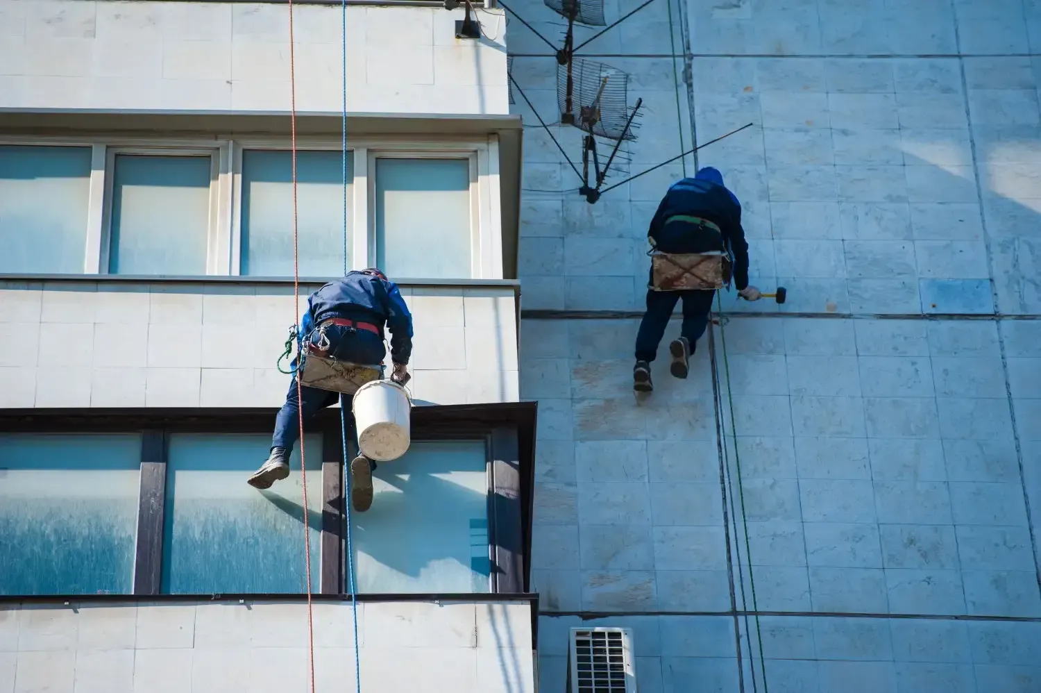 Dos trabajadores, sujetos con cuerdas, limpian el exterior de un edificio de apartamentos azul con cubos y herramientas.