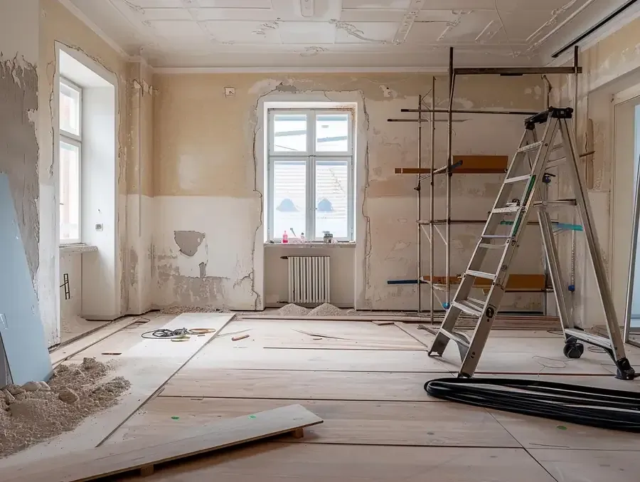 Obras de renovación en curso en una habitación luminosa con escalera, andamios, herramientas y paredes y suelo sin terminar.