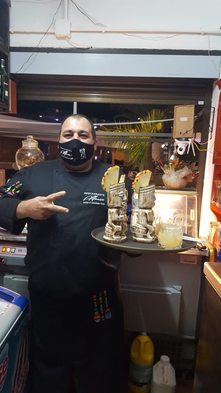 Hombre sosteniendo una bandeja con bebidas y comida, usando una máscara y un delantal, haciendo un signo de paz.