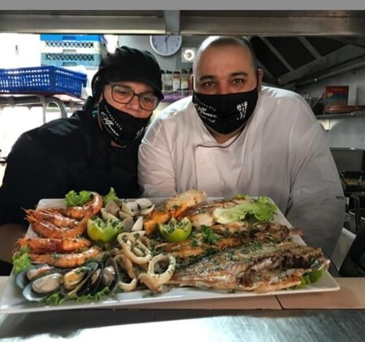 Dos personas con máscaras sosteniendo una bandeja de mariscos cocidos en una cocina.