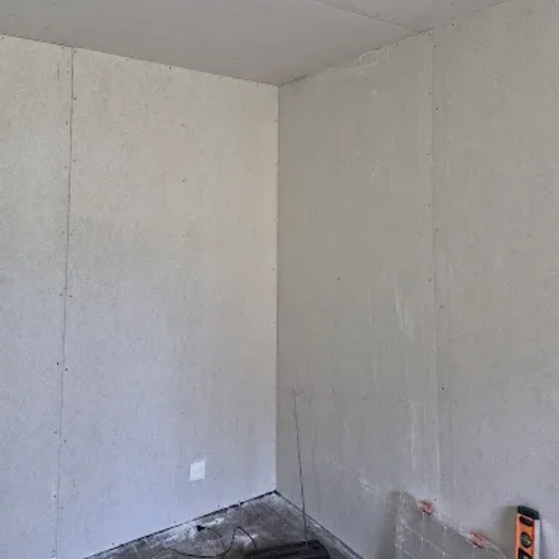 Coin de pièce blanc nu avec murs non finis, sol apparent et une petite prise électrique murale.