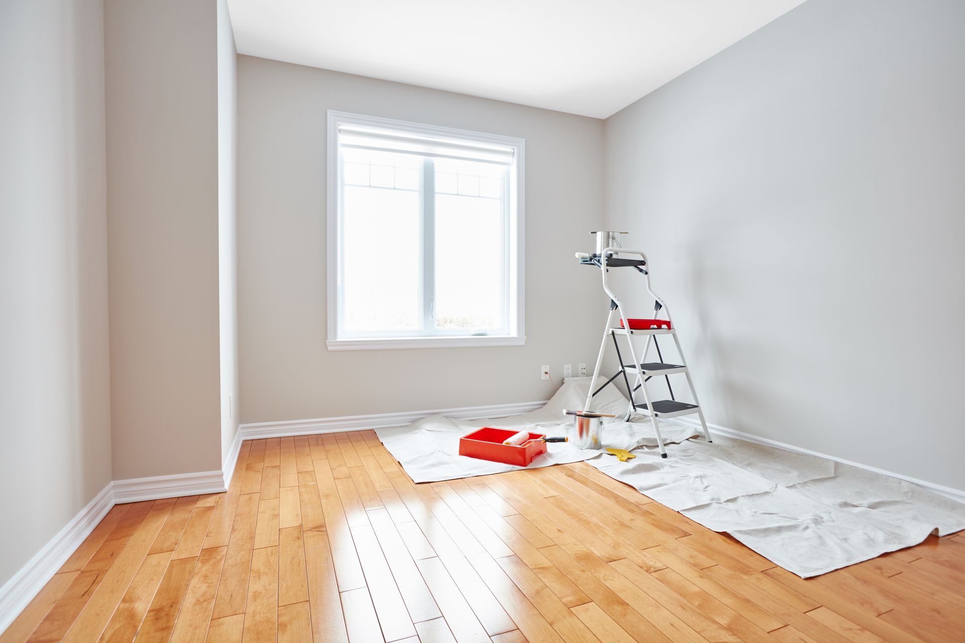 Pièce lumineuse avec parquet, une fenêtre et du matériel de peinture sur une bâche.