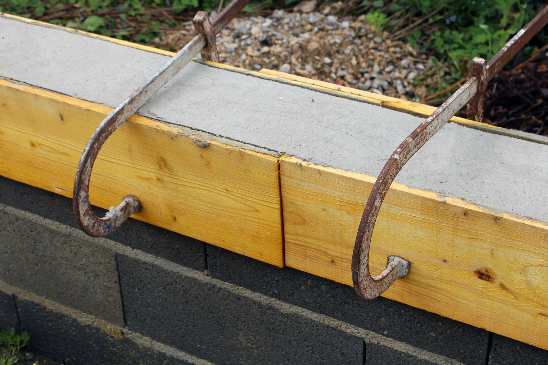 Des crochets en fer à béton rouillés fixent une planche de bois jaune à un mur en parpaings.