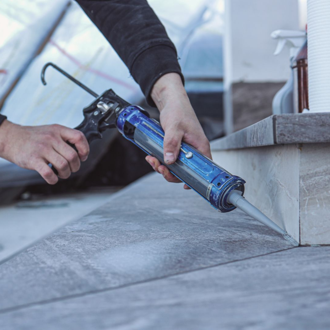 Des mains utilisent un pistolet à calfeutrer bleu pour sceller un joint de béton le long d'un mur.